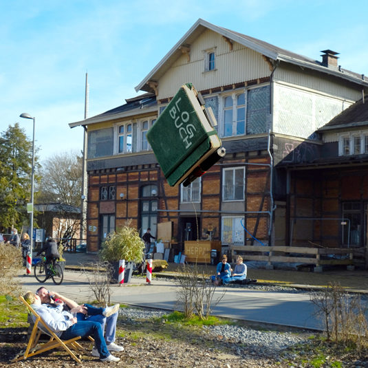 Utopiastadt-Campus-Flächen mit Blick auf den Nordost-Giebel des Mirker Bahnhofs. Davor 'fliegt' ein offensichtlich hochgeworfenes Notizbuch mit der Aufschrift »LOGBUCH«.