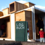 Auf einer Beton-Kante steht ein Notizbuch mit der Aufschrift »LOGBUCH«. Im Hintergrund sieht man unscharf Kinderspielcontainer.
