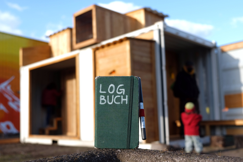 Auf einer Beton-Kante steht ein Notizbuch mit der Aufschrift »LOGBUCH«. Im Hintergrund sieht man unscharf Kinderspielcontainer.