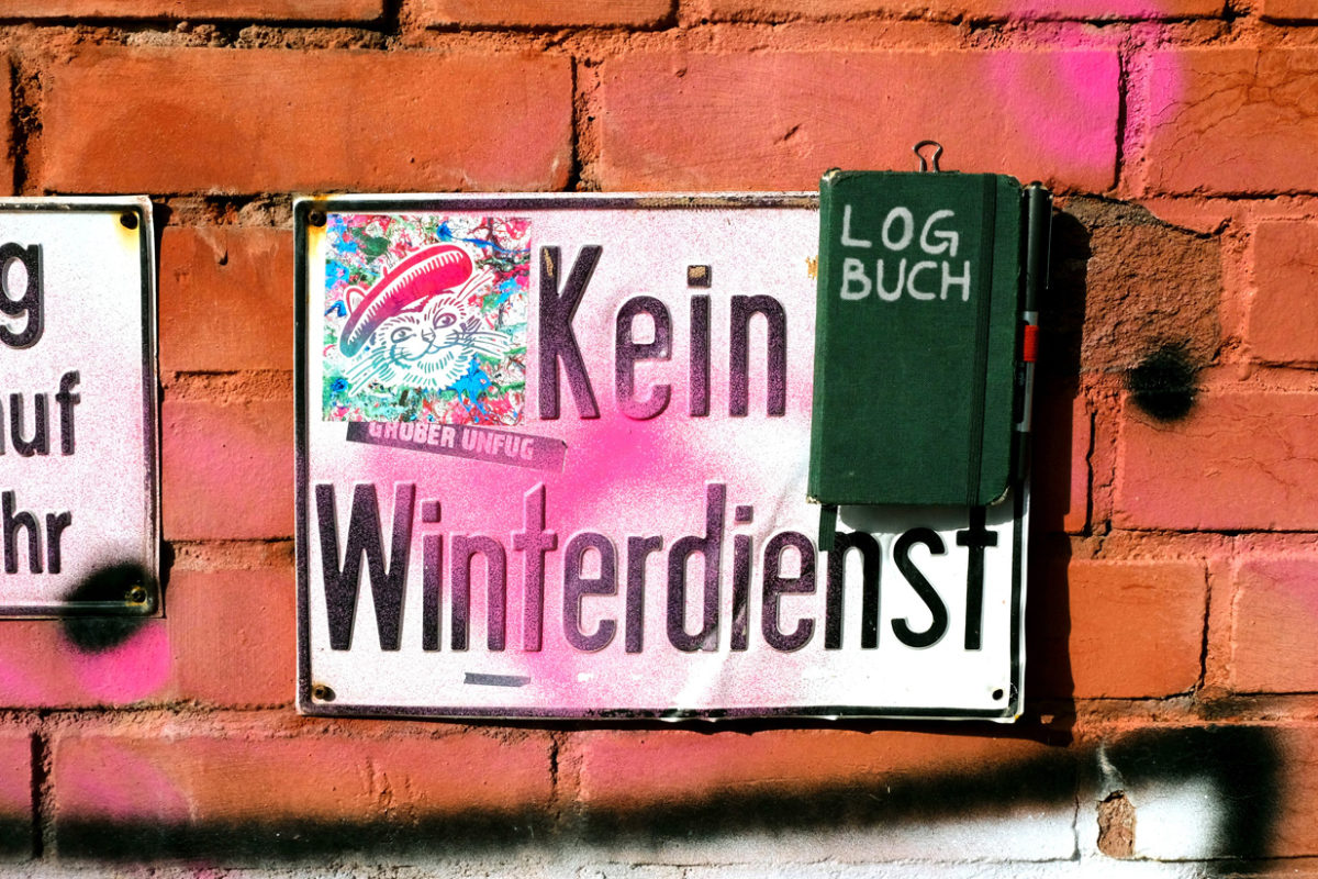 An einer angemalten Backstein-Wand hängt ein besprühtes und verbeultes Schild mit der Aufschrift »Kein Winterdienst« und an diesem Schild hängt ein Notizbuch mit der Aufschrift »LOGBUCH«