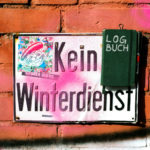 An einer angemalten Backstein-Wand hängt ein besprühtes und verbeultes Schild mit der Aufschrift »Kein Winterdienst« und an diesem Schild hängt ein Notizbuch mit der Aufschrift »LOGBUCH«