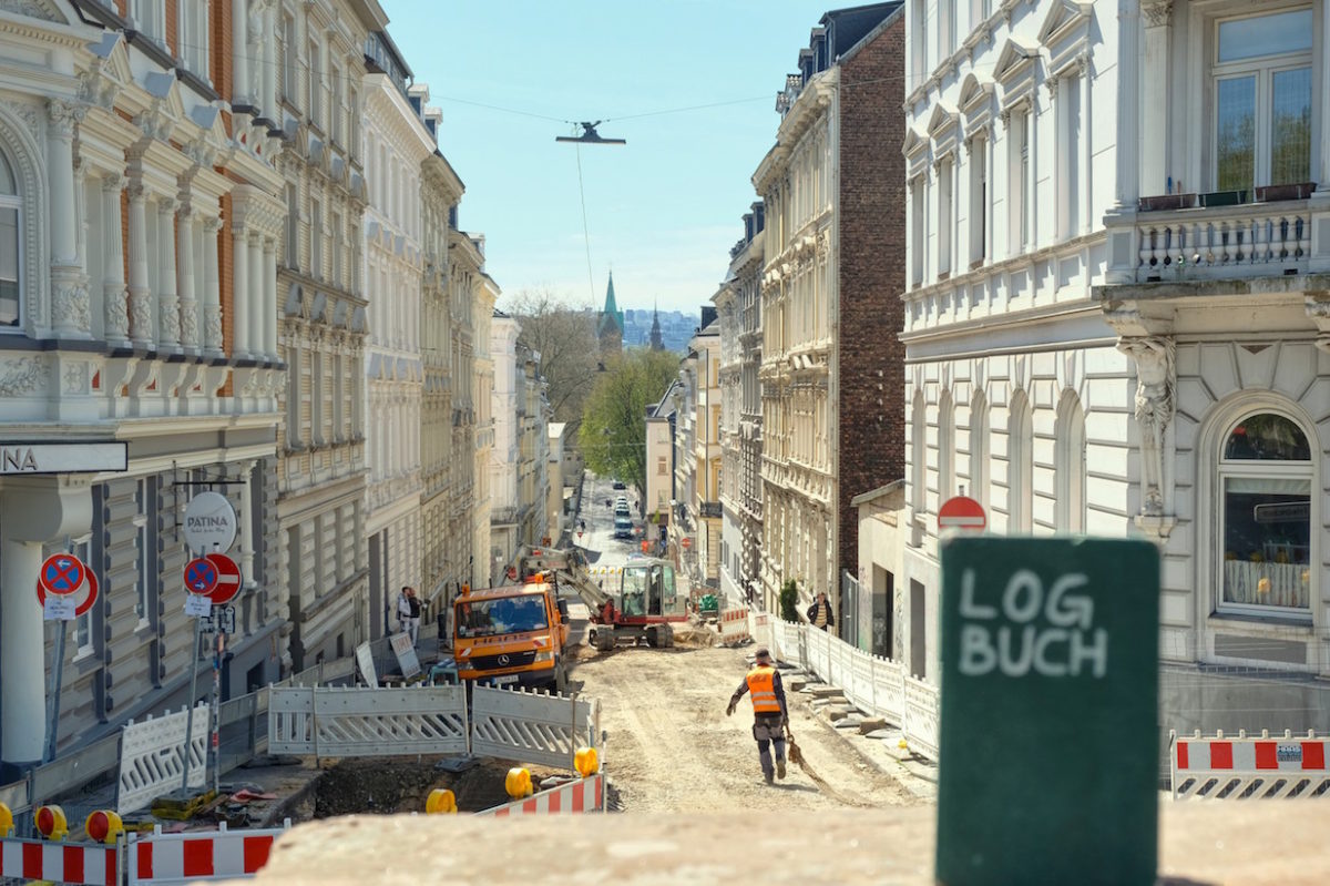 Blick auf die Neue Norsdstraße, die komplette Baustelle ist. Im Vordergrund steht leicht unscharf ein Notizbuch mit der Aufschrift »LOGBUCH« auf dem Mauer-Sims.