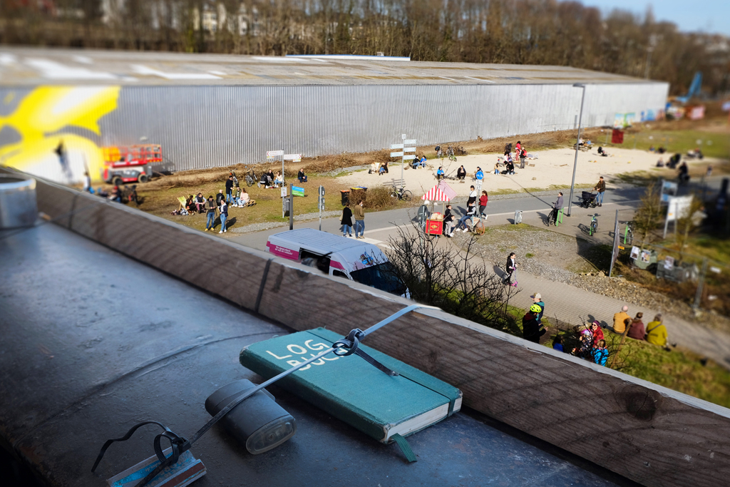 Blick vom Balkon auf den Utopiastadt-Campus. Auf dem Balkonsims liegt ein Notizbuch mit der Aufschrift »LOGBUCH«