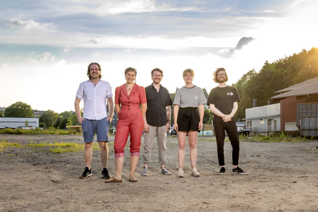 Das Bild zeigt 5 Personen (Achim, Amanda, David, Clara und Mosche) die auf der Freifläche des Utopiastadt Campus stehen
