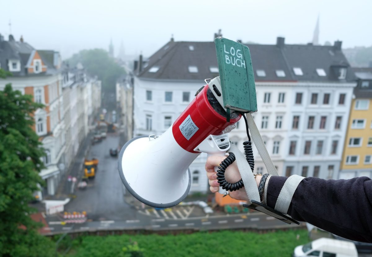 Ein Megaphon ist aus großer Höhe Richtung Quartier gerichtet. An der Sprecheinheit hängt ein Notizbuch mit der Aufschrift »LOGBUCH«