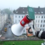 Ein Megaphon ist aus großer Höhe Richtung Quartier gerichtet. An der Sprecheinheit hängt ein Notizbuch mit der Aufschrift »LOGBUCH«