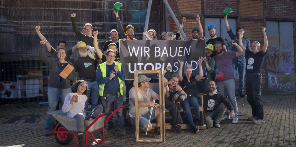 Viele fröhliche Utopist:innen mit Helmen, Handschuhen, Warnwesten, Werkzeug und Baumaterialien halten in der Mitte ein Banner mit der Aufschrift »Wir bauen Utopiastadt«.