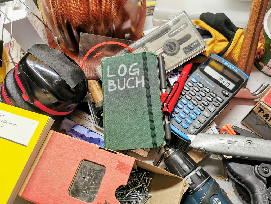 Ein Tisch voller Werkzeuge, mittendrin ein Notizbuch mit der Aufschrift »LOGBUCH«