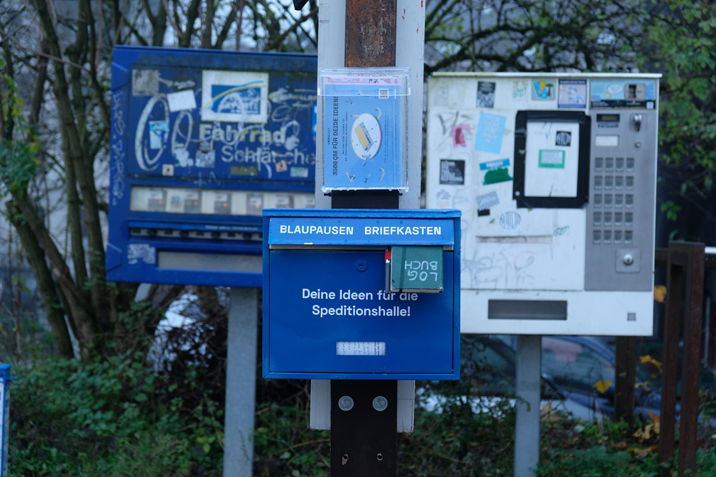 Blauer Briefkasten mit Aufschrift »Deine Ideen für die Speditionshalle!«, im Briefschlitz steckt zur hälfte ein Notizbuch mit der Aufschrif »LOGBUCH«.