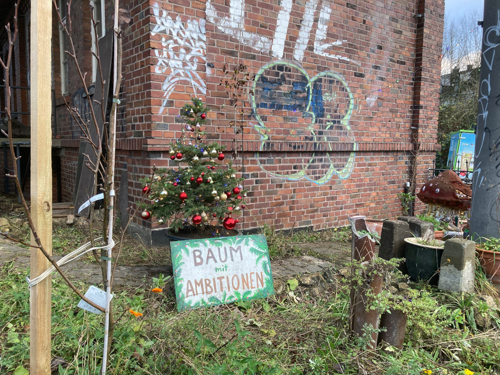 Geschmückter Nadelbaum vor Klinker-Wand der GPA, an dem ein Schild lehnt: »Baum mit Ambitionen«.