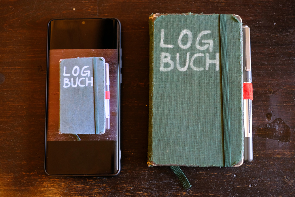 Ein Smartphone, auf dessen Bildschirm das Foto eines Notizbuches mit der Aufschrift »LOGBUCH« zu sehen ist liegt neben einem Notizbuch mit der Aufschrift »LOGBUCH«