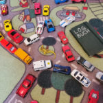 Spielzeugteppich mit Straßendekor, vollgestellt mit Spielautos. Daneben liegt ein Notitzbuch mit der Aufschrift »LOGBUCH«.