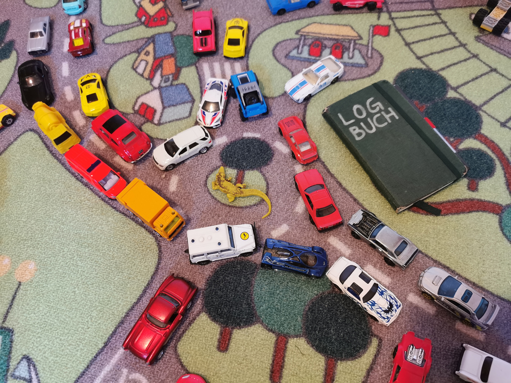 Spielzeugteppich mit Straßendekor, vollgestellt mit Spielautos. Daneben liegt ein Notitzbuch mit der Aufschrift »LOGBUCH«.