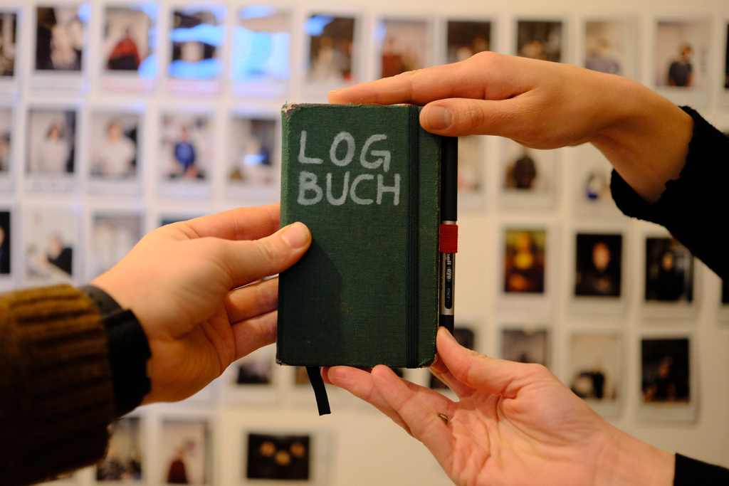 Drei unterschiedliche Hände halten ein Notizbuch mit der Aufschrift LOGBUCH hoch. Im unscharfen Hintergrund hängen zahlreiche Polaroid-Portraits an der Wand.