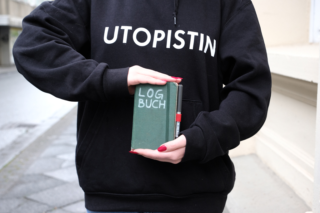 Person mit rot lackierten Fingernägeln und Pulli, auf dem »Utopistin« steht, hält ein Notizbuch mit der Aufschrift »LOGBUCH«. Man sieht nur diesen Ausschnitt. Kein Gesicht, keine Beine.