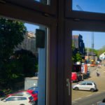 Blick aus dem Fenster auf Bäume vor dem Verhüllten Bahnhofsgebäude. Außerdem lebhafter Verkehr auf dem Bahnhofsvorplatz zwischen Bauzäunen und Kran. An der Fensterscheibe hängt ein Notizbuch mit der Aufschift LOGBUCH.