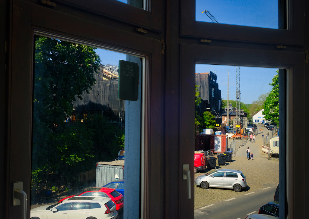 Blick aus dem Fenster auf Bäume vor dem Verhüllten Bahnhofsgebäude. Außerdem lebhafter Verkehr auf dem Bahnhofsvorplatz zwischen Bauzäunen und Kran. An der Fensterscheibe hängt ein Notizbuch mit der Aufschift LOGBUCH.