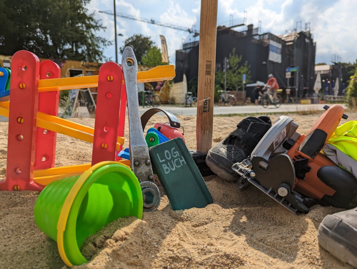 In einem Sandkasten liegen Handwerkszeug und Kinderspielzeug munter nebeneinander und mittendrin steckt ein Notizbuch mit der Aufschrift »LOGBUCH«.