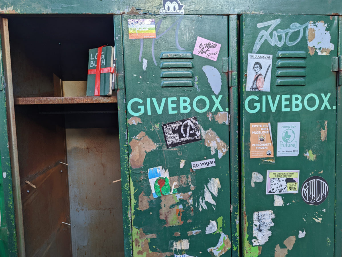 Runtergerockter 3er-Spind mit Aufschrift 'GIVEBOX'. Auf einem Regalbrett steht ein mit Geschenkband versehenes Notizbuch, auf dem 'LOGBUCH' steht.