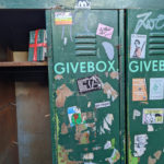 Runtergerockter 3er-Spind mit Aufschrift 'GIVEBOX'. Auf einem Regalbrett steht ein mit Geschenkband versehenes Notizbuch, auf dem 'LOGBUCH' steht.