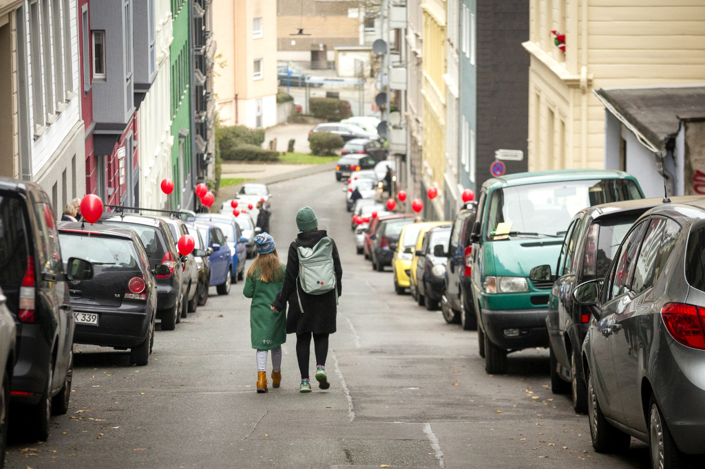 Zwei Kinder gehen Hand in Hand eine Straße herunter, rechts und links parken überall Autos, an vielen davon sind rote Luftballons befestigt