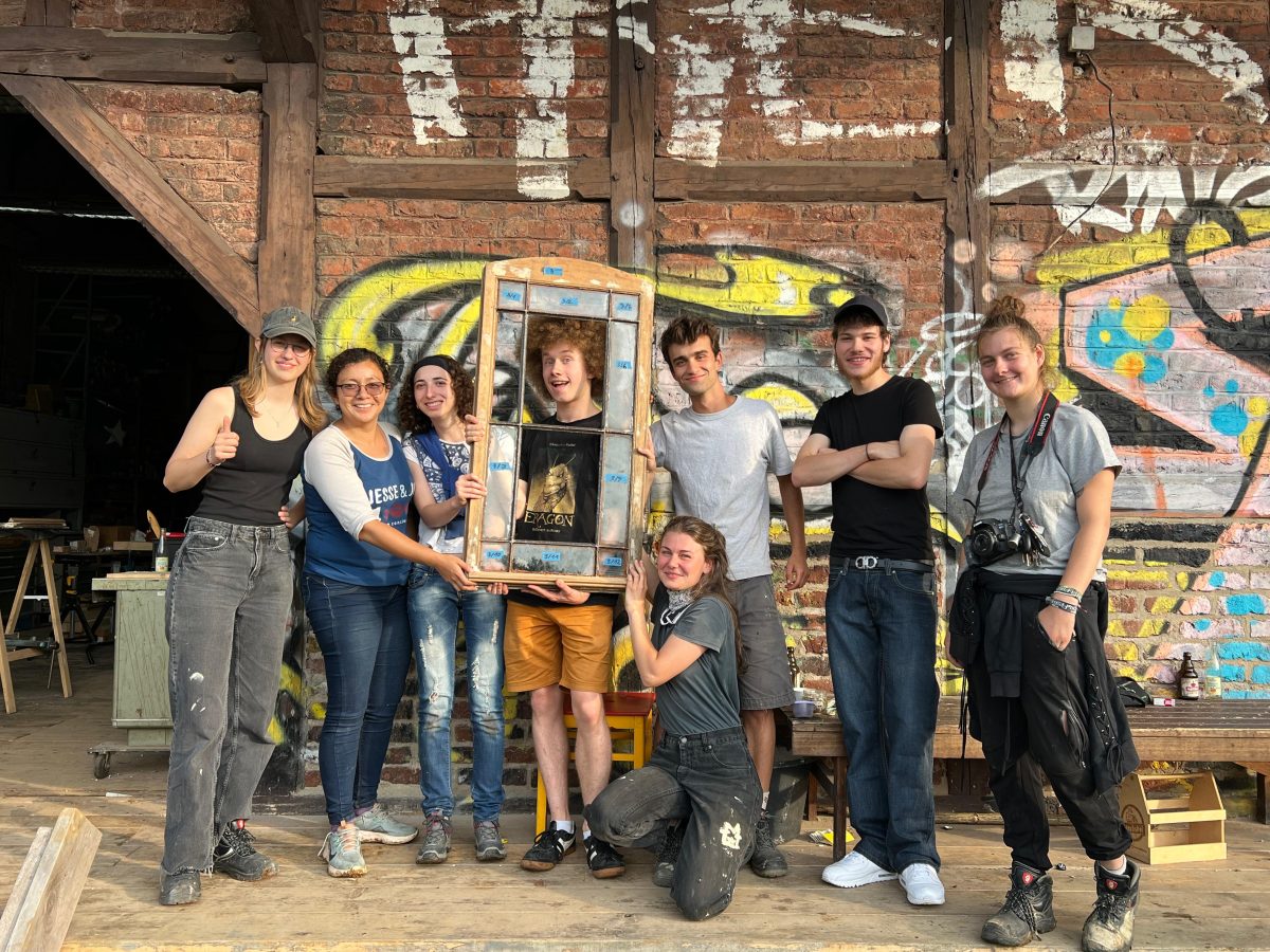 Junge Menschen stehen vor einer mit Grafitti verzierten Fachwerk-Wand und lachen fröhlich in die Kamera, einer hält ein historisches Fenster im Sanierungszustand hoch und lacht hindurch.